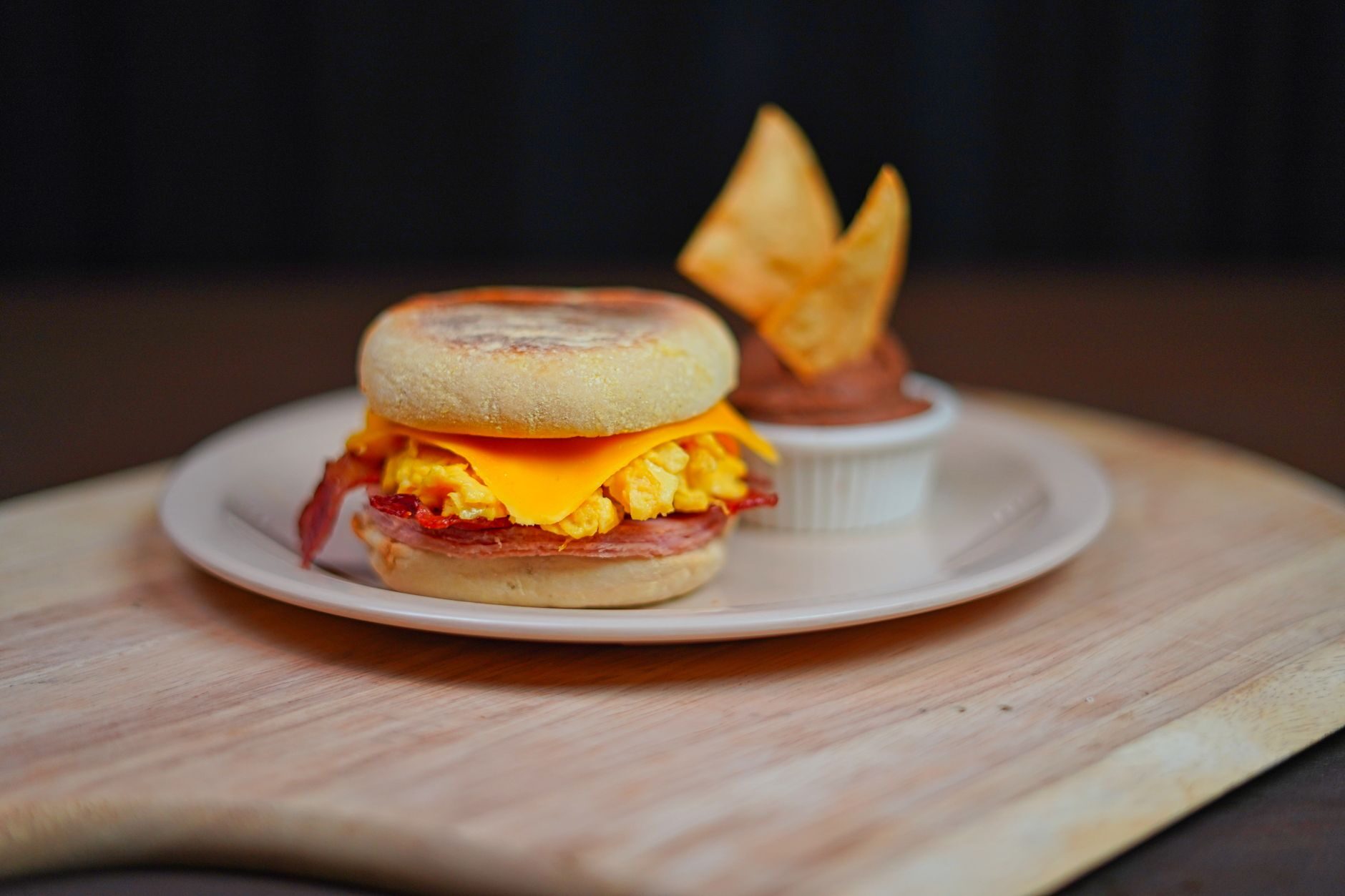An egg, bacon and melting cheese breakfast sandwich on a muffin with a cup of dipping sauce