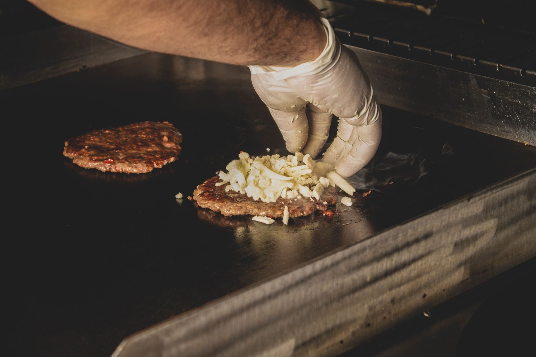 A white-gloved hand pressing patties on the flat-top grill with grilled onions