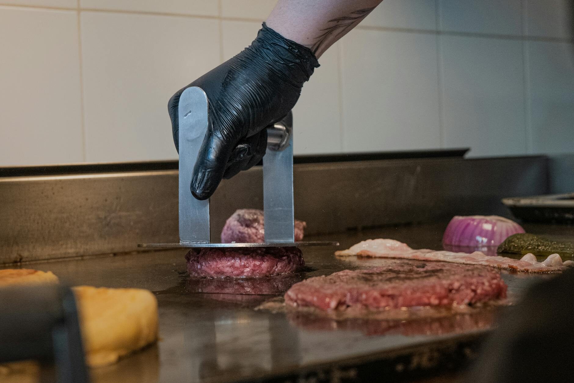 A gloved hand pressing a smash tool onto a beef patty on the flat-top grill beside grilled onions