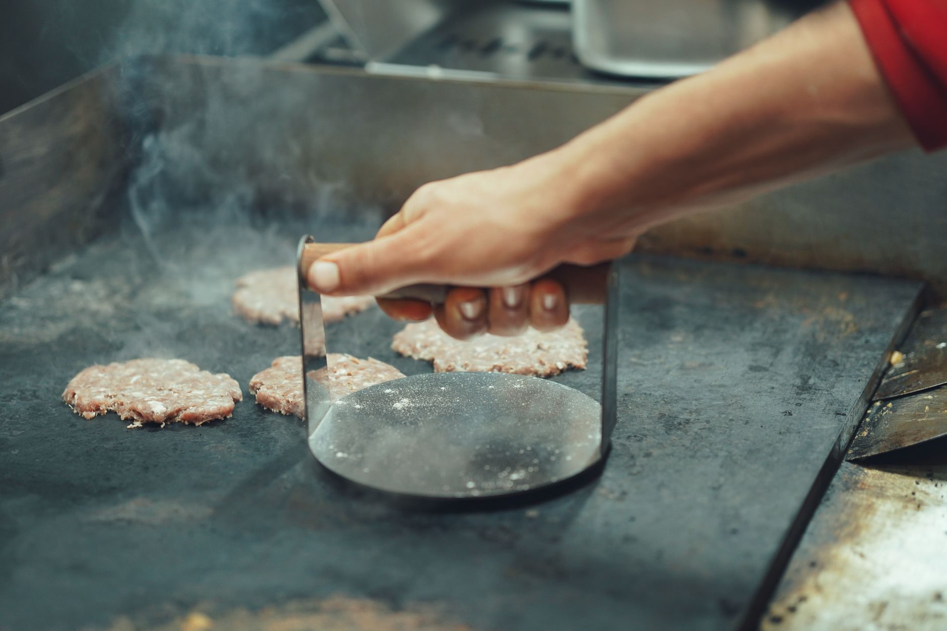 Cook smashing burger patties on a steaming commercial flat-top griddle with a burger press