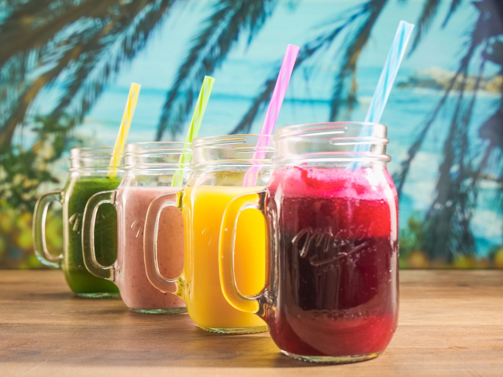Four colorful fruit smoothies in mason jars with straws.