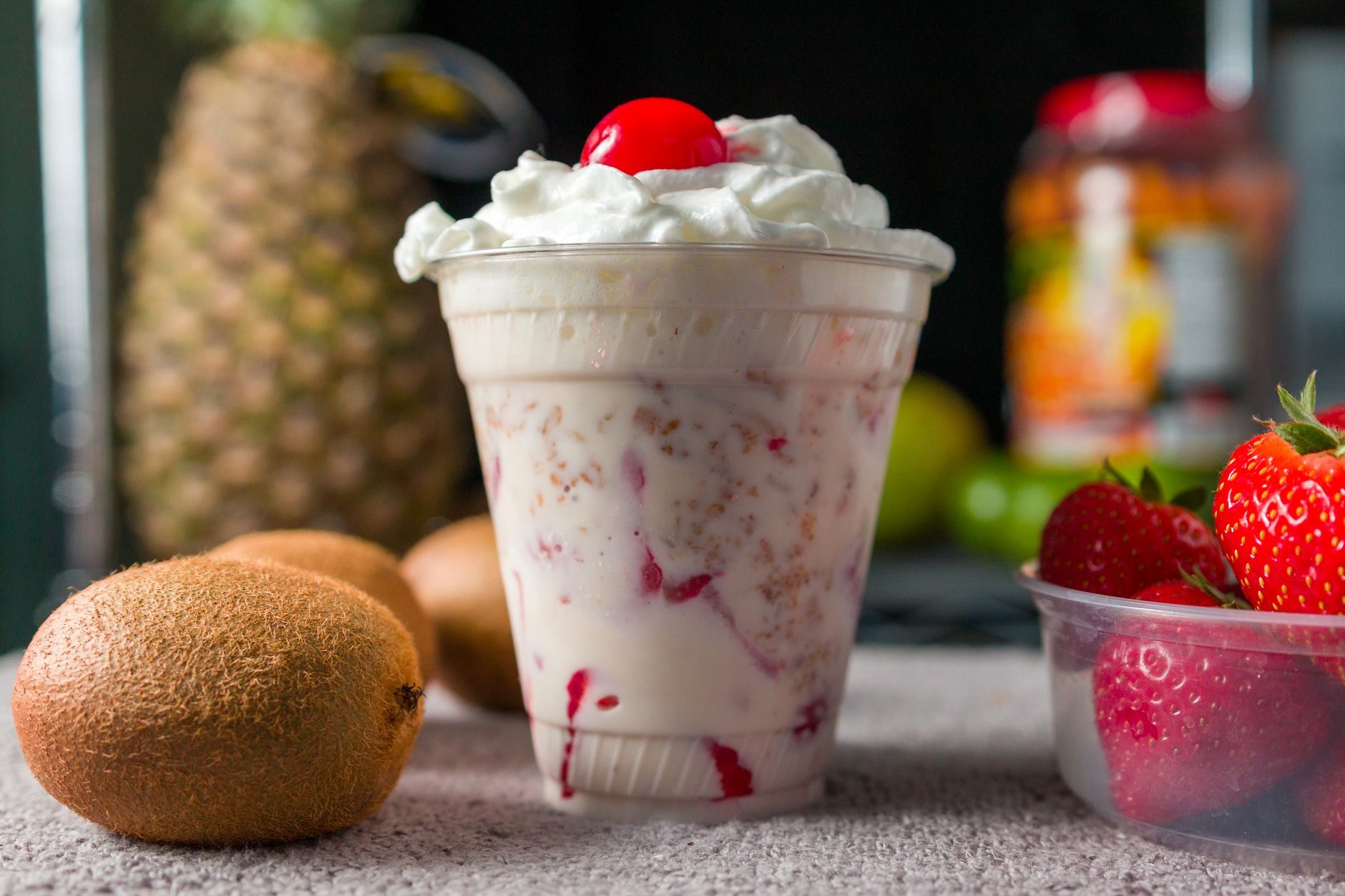 A strawberry smoothie in a cup surrounded by fresh pineapple, kiwi and strawberries